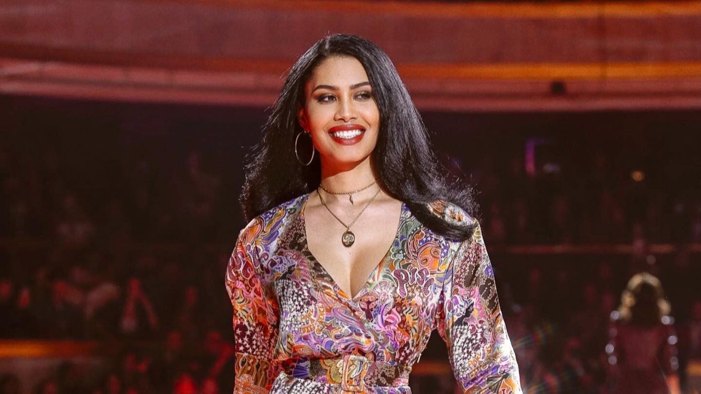 Leyna Bloom confidently struts down a brightly lit runway in a colorful patterned dress, smiling as the audience watches.