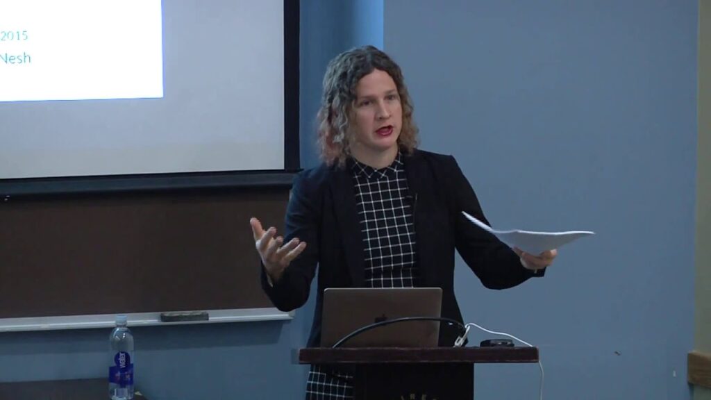 Dr. micha cárdenas presents her research on trans embodiment and digital art while standing at a podium with laptop and notes.