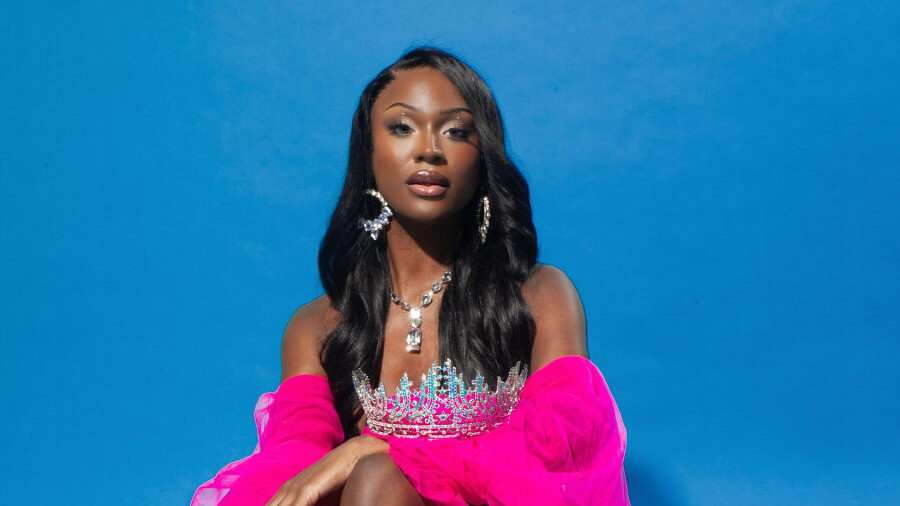 Midori Monet, wearing a bright pink gown and crown, poses seated in front of a blue background with flowers.