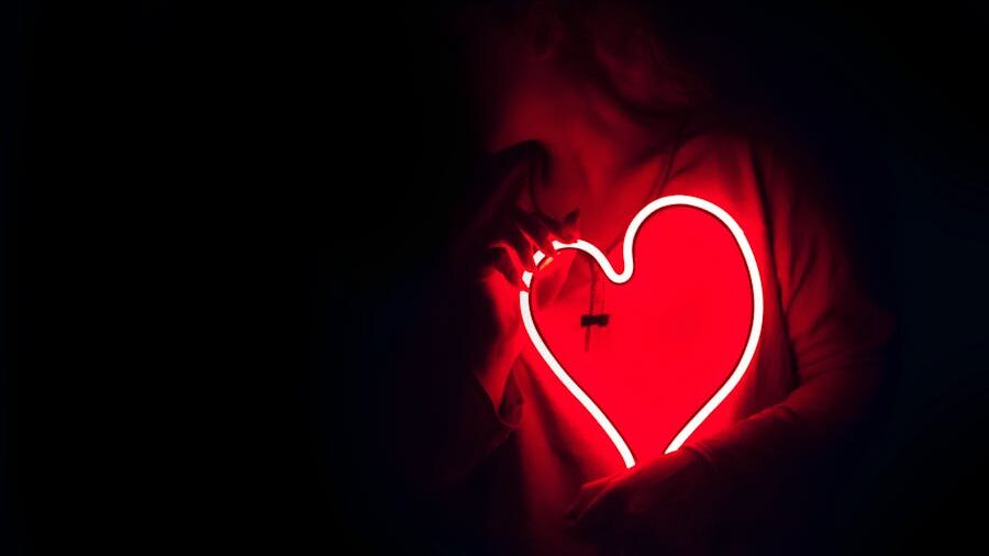 Person holding a glowing red neon heart in the dark, with the light illuminating their chest.