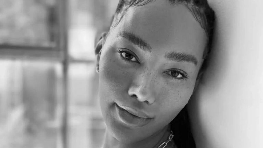 Black and white portrait of Munroe Bergdorf leaning on a window frame, makeup-free, wearing a printed top and chain necklace.