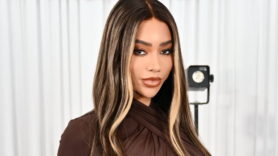 Glamorous portrait of Munroe Bergdorf with long highlighted hair and a ruched brown dress in a studio setting.