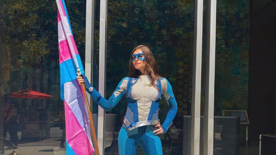 Nicole Maines con disfraz de superheroína, sosteniendo una gran bandera trans frente a un edificio moderno de vidrio.