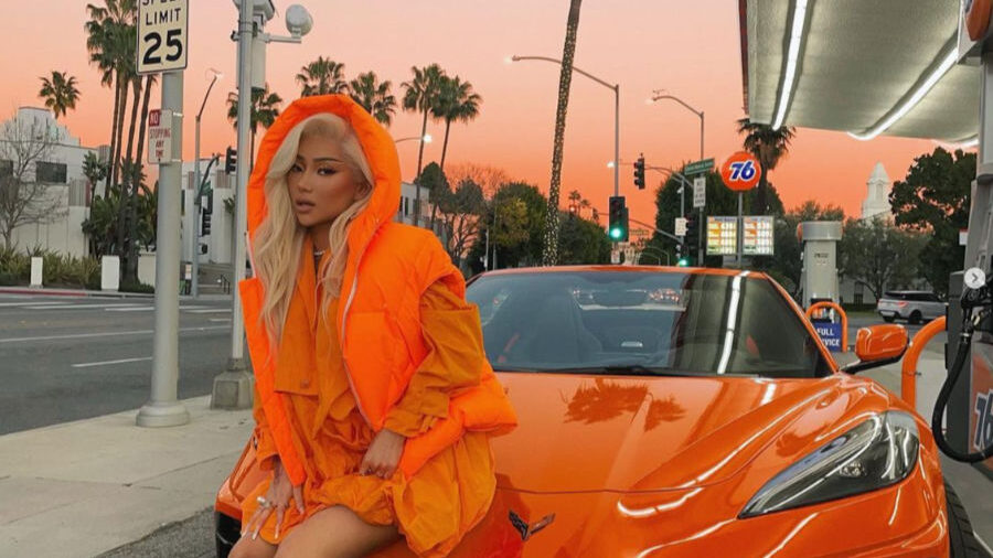 Nikita Dragun in bright orange outfit sitting on the hood of a matching Corvette.