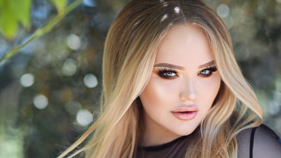 NikkieTutorials con el cabello rubio largo, usando un top negro de malla bordado con flores, en una sesión al aire libre.