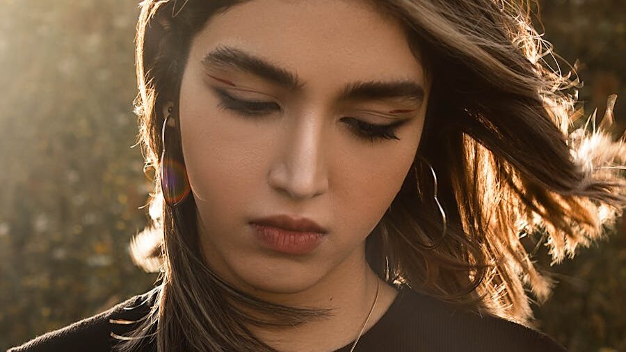 young woman with shoulder-length hair and cross necklace looks down, bathed in soft sunlight, flowers blurred in front.