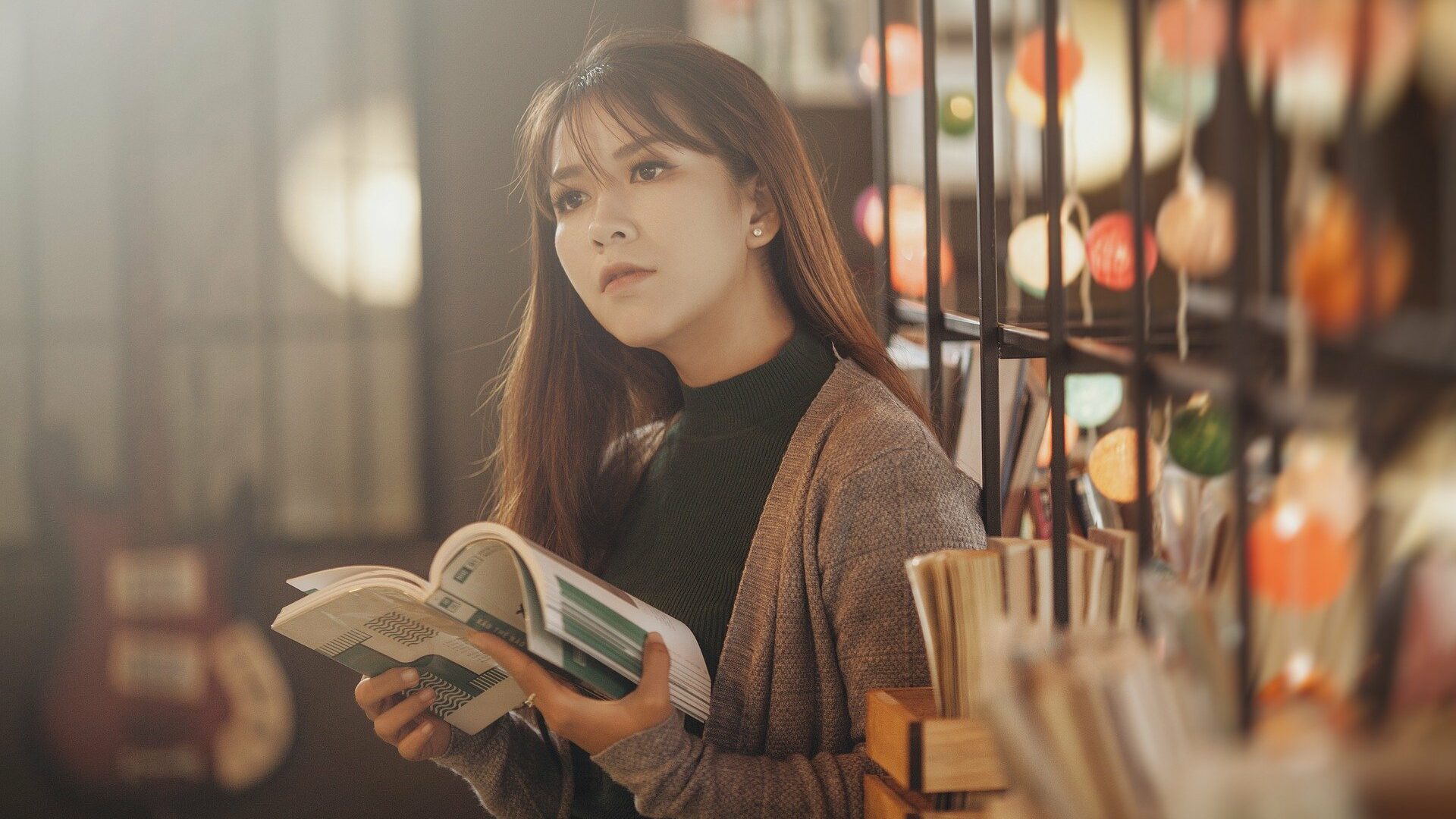 Woman with long brown hair reading a book near a decorated bookshelf in a softly lit library.