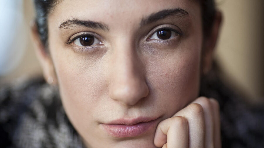 Close-up portrait of a young woman with dark hair, resting her chin on her hand, gazing directly into the camera.