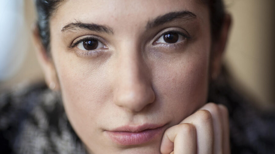 Retrato en primer plano de una mujer joven de cabello oscuro que mira directamente a la cámara y tiene la barbilla apoyada en la mano.