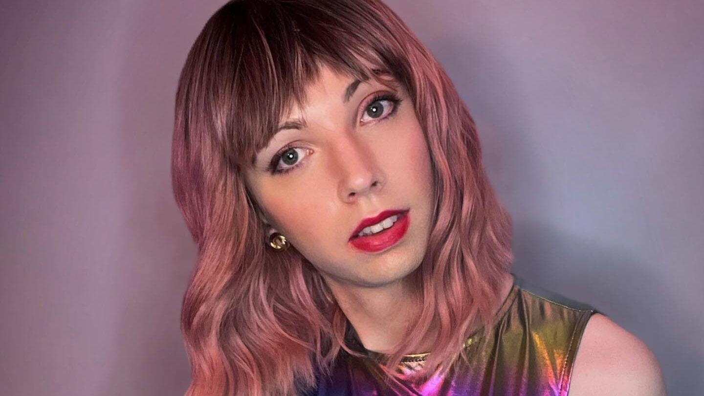 Rosie, con un brillante vestido iridiscente de arco iris, el cabello rosa ondulado hasta los hombros y una postura segura contra un fondo neutro.