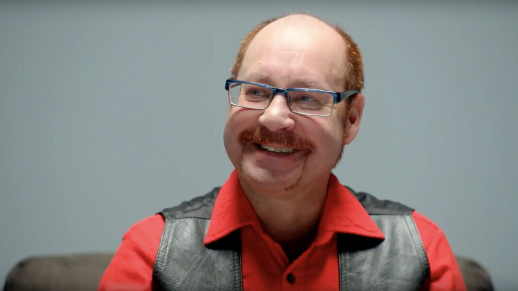 Rupert Raj, wearing glasses and a leather vest, speaks during an interview on a gray background.