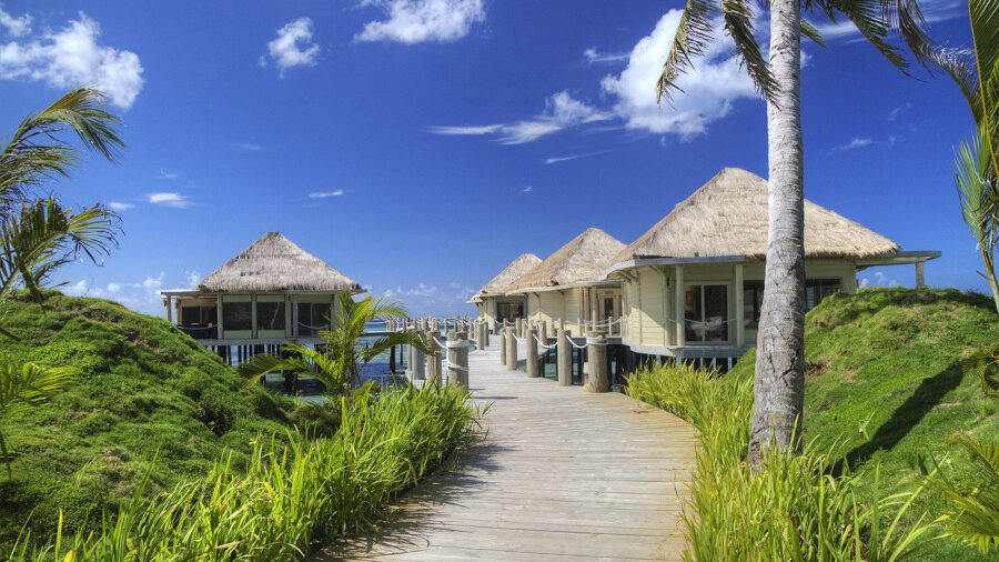 Station balnéaire avec palmiers et bungalows sur pilotis sous un ciel bleu vif parsemé de nuages.