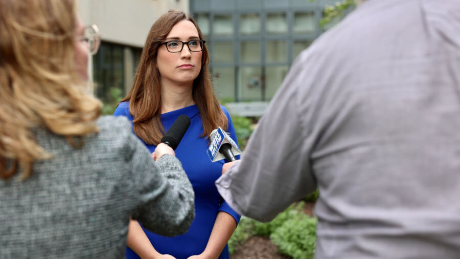 Sarah McBride in a blue dress speaking to journalists holding microphones.