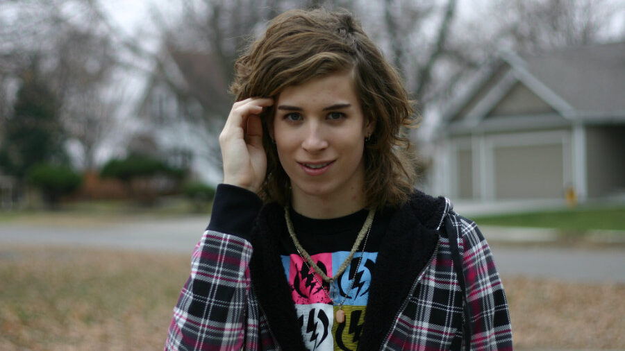 Aimee Ardell de pie afuera, con una chaqueta a cuadros sobre una camiseta colorida, con el pelo ondulado hasta los hombros y una sonrisa llena de confianza.