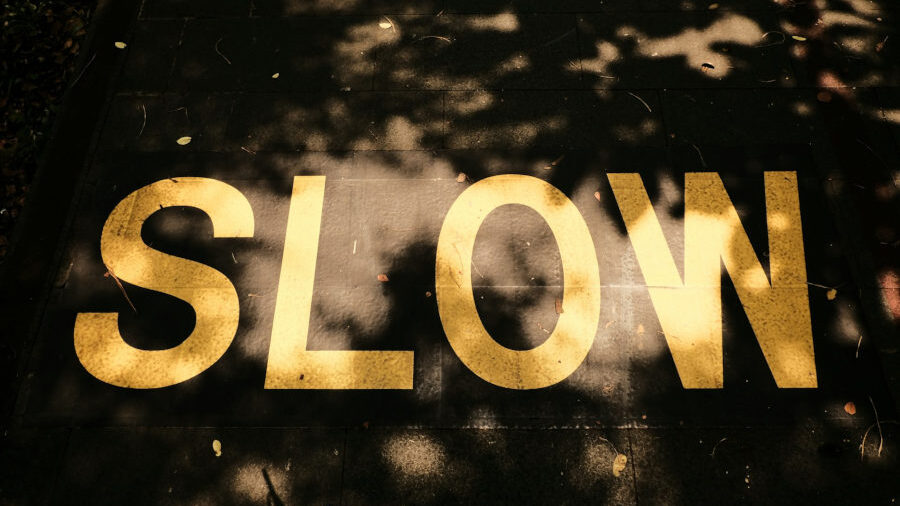 Large yellow “SLOW” painted on dark stone pavement, partially covered by sunlight filtering through trees.