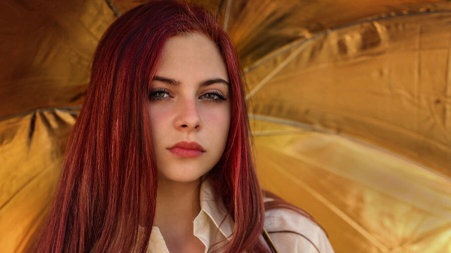 Mujer joven con cabello largo y rojo, de pie frente a un fondo dorado.