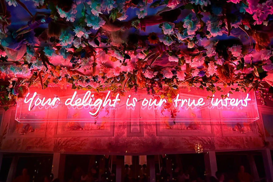 Neon sign “Your delight is our true intent” under a ceiling filled with Barbies and artificial flowers.