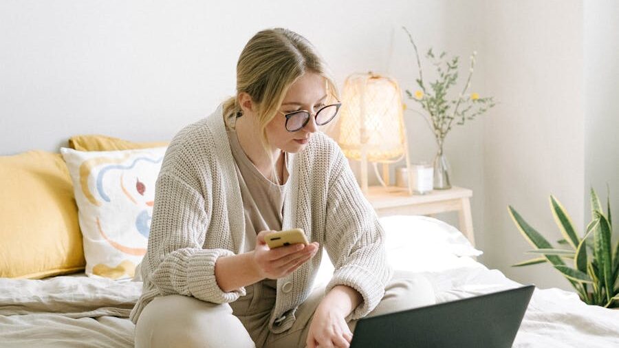 Woman with glasses sitting on bed, checking her phone and laptop while reviewing an online profile in a softly lit bedroom.