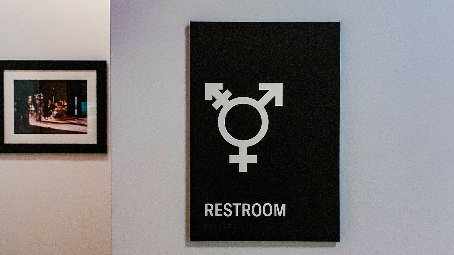 A black sign with a white transgender symbol and the word “Restroom” in bold text, mounted on a wall next to a framed picture.