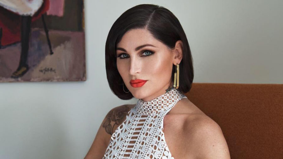 Trace Lysette sentada en una silla naranja, con un vestido blanco sin mangas de ganchillo.