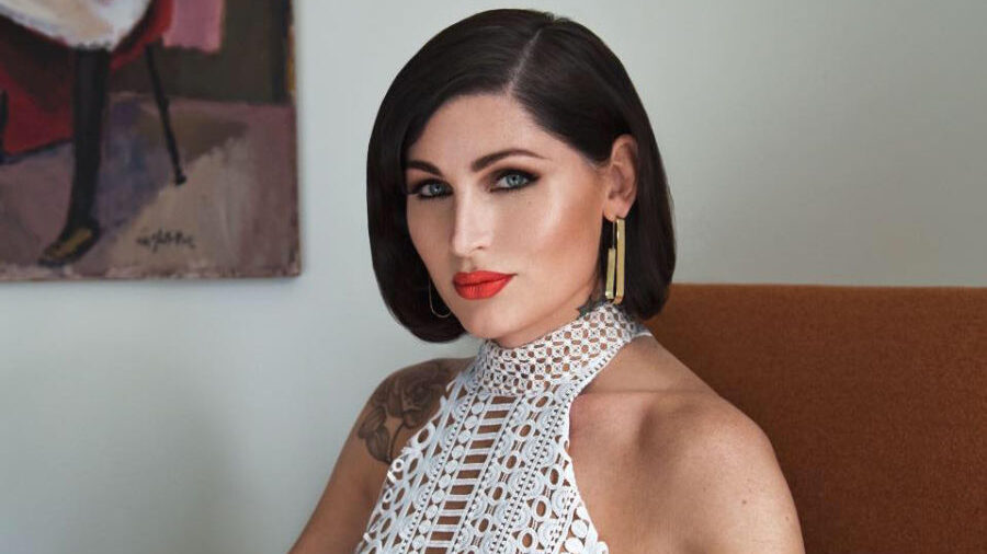 Trace Lysette seated on an orange chair, wearing a sleeveless white crochet dress.