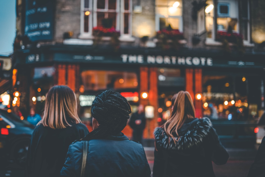 Three trans women seen from behind standing outside The Northcote pub in London, lit warmly in the evening.