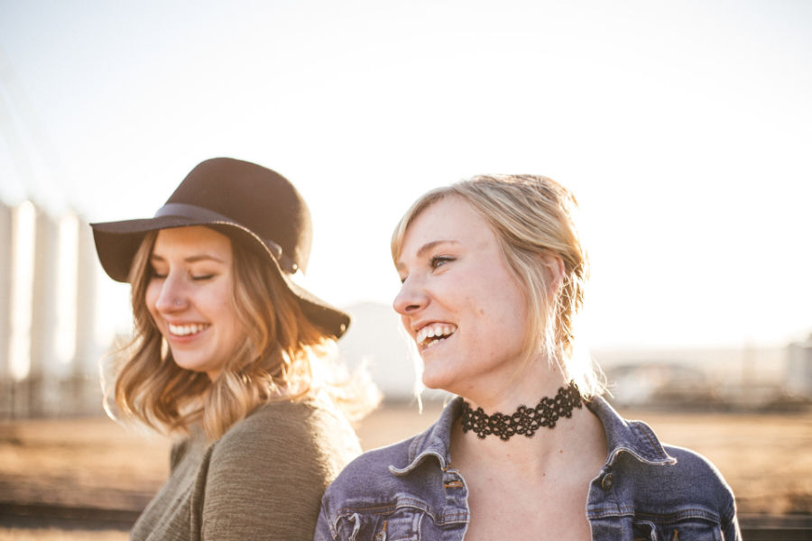 Two trans women smiling and laughing together in soft sunlight, one wearing a black hat and the other with a choker necklace.