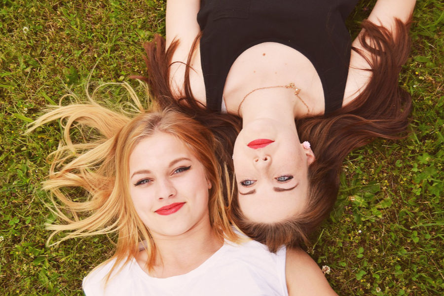 A trans woman from Bakersfield and her friend lying on green grass, smiling and facing the camera in a peaceful setting.