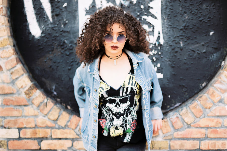 Trans woman with curly hair, sunglasses, choker, and denim jacket standing in front of a round black wall.