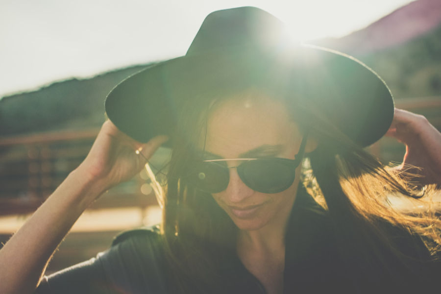 A trans woman wearing sunglasses and a hat in sunlight, photographed outdoors in Denver.