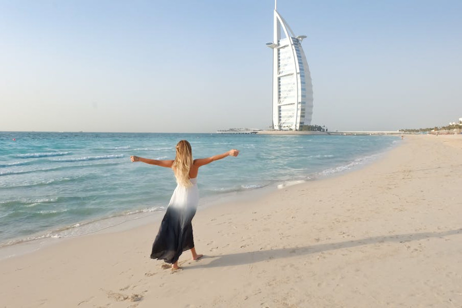 Trans woman in a white and black dress standing barefoot on a Dubai beach, facing the sea, with Burj Al Arab visible in background.