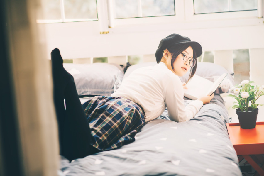 Trans woman with glasses and a hat lying on a bed, holding a book, next to a potted flower on a red table by the window.