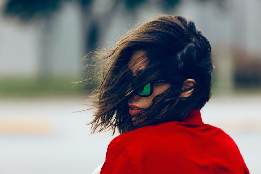 Trans woman wearing a red jacket and black sunglasses, hair blown by the wind, facing sideways.