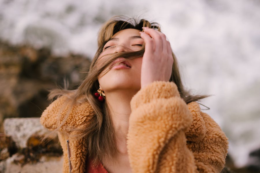 A trans woman in a tan coat outdoors with eyes closed and hair gently covering her face, expressing calm and confidence.