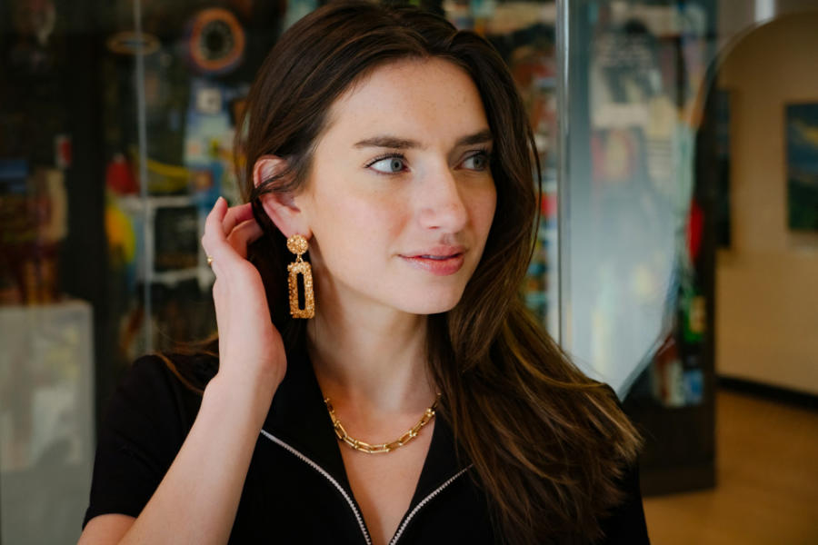 Trans woman with long brown hair wearing large gold earrings and a chain necklace, adjusting her earring in a public indoor space.