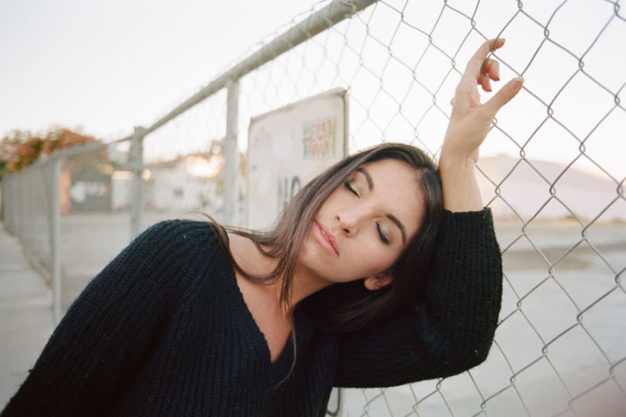Trans woman in black sweater leaning on a chain-link fence, eyes closed, in soft San Diego evening light.