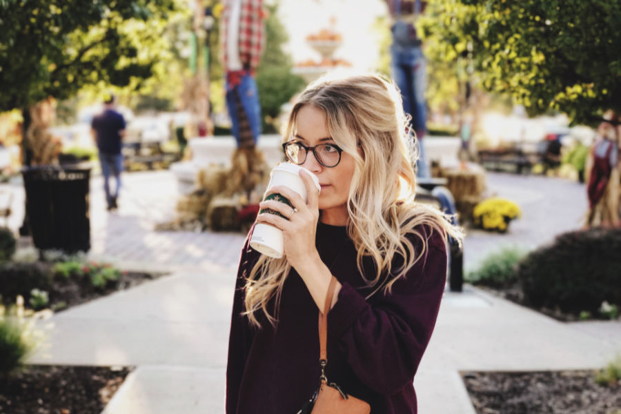 A trans woman with long blonde hair and glasses drinks coffee outside in San Jose on a sunny autumn day.