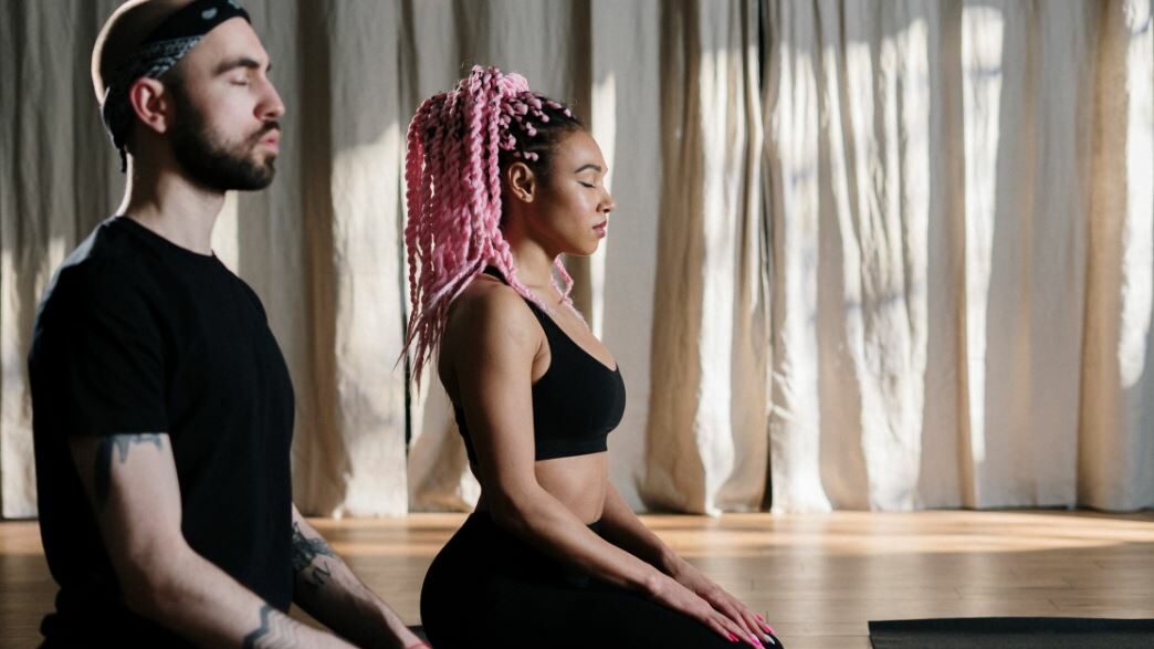 Femme aux tresses roses et homme avec un bandeau méditant côte à côte.