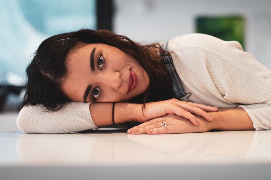 Trans woman resting her head on a white table, wearing a light sweater and looking softly at the camera in a calm, indoor setting.