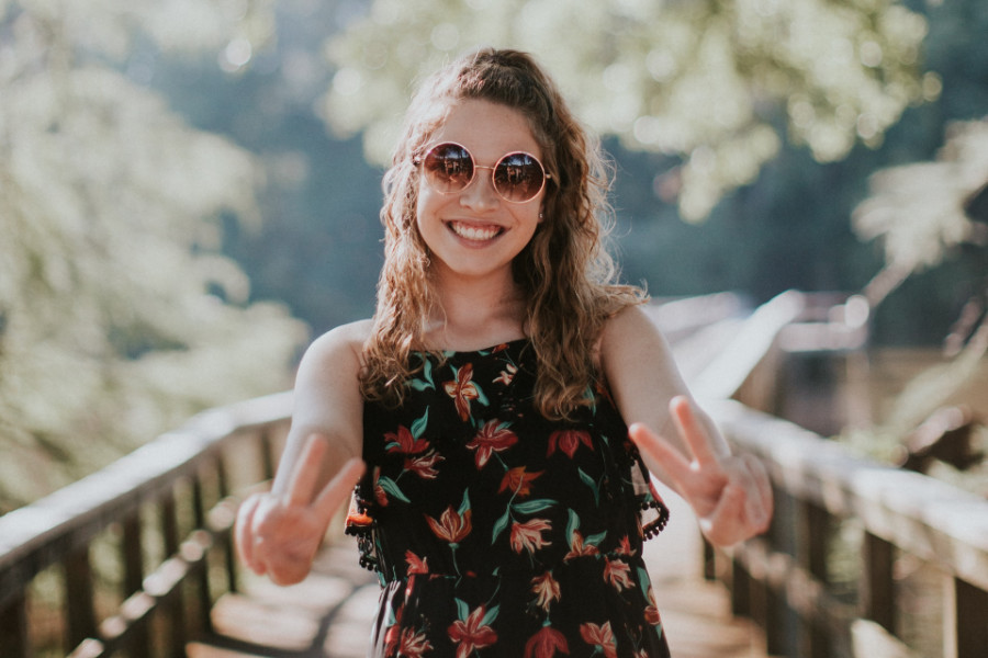 A trans woman from Miami smiles brightly while making peace signs with both hands on a sunlit wooden bridge.