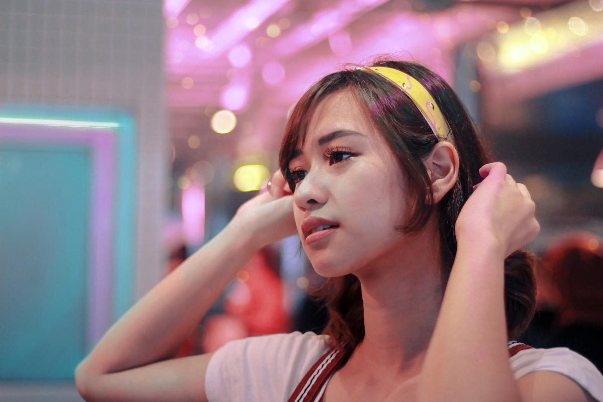 Close-up of a trans woman in a white shirt and headband, set against a colorful background with dreamy lighting.