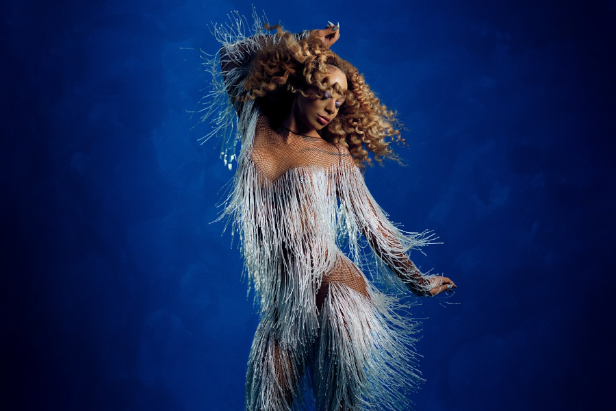 Urias in a glittery silver fringe bodysuit, mid-dance pose, on a blue studio set.