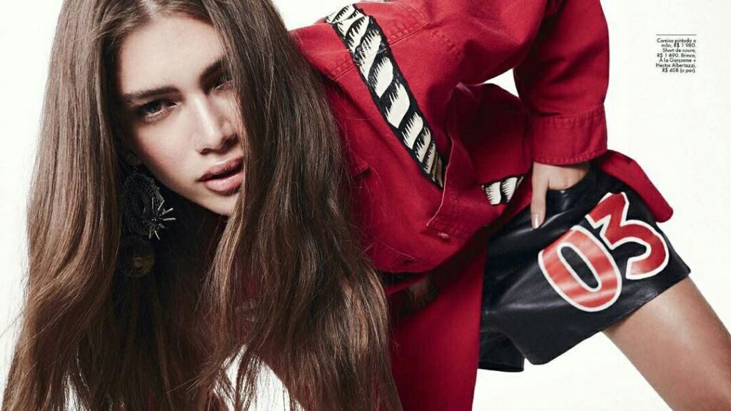 Valentina Sampaio con top rojo y shorts negros, inclinada hacia adelante durante una sesión de fotos de moda.