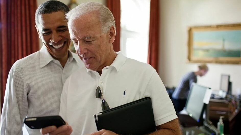 Joe Biden et Obama souriant en regardant un smartphone, debout dans un bureau.