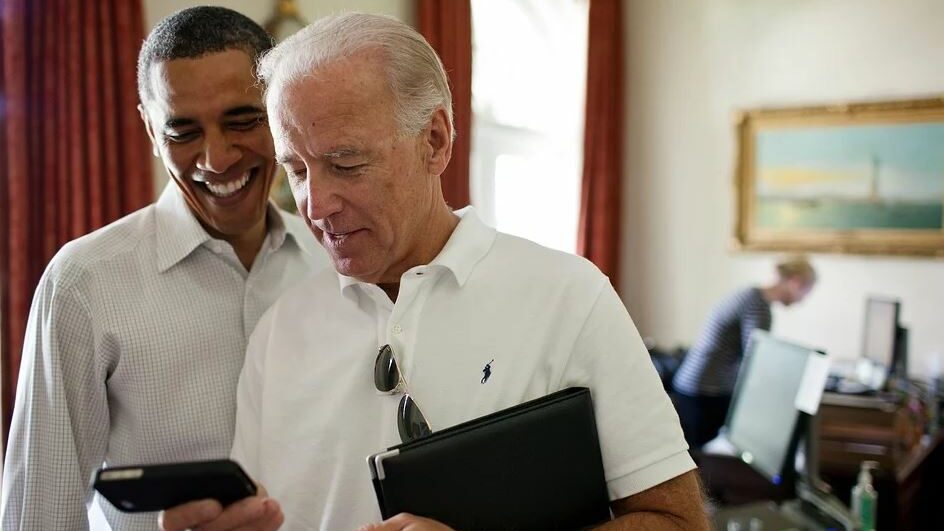 Joe Biden and Obama smiling while looking at a smartphone, standing in an office.