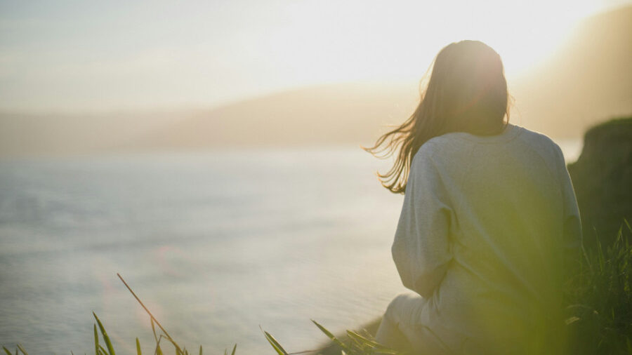 Persona sentada en una colina con hierba mirando el agua al atardecer.